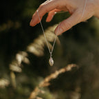 A delicate silver necklace with a small feather pendant is held by a hand against a blurred outdoor background of green foliage and warm sunlight.