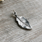 A sterling silver feather pendant with detailed textured lines and a '925' silver purity stamp and hallmark, resting on a rustic grey wood surface.