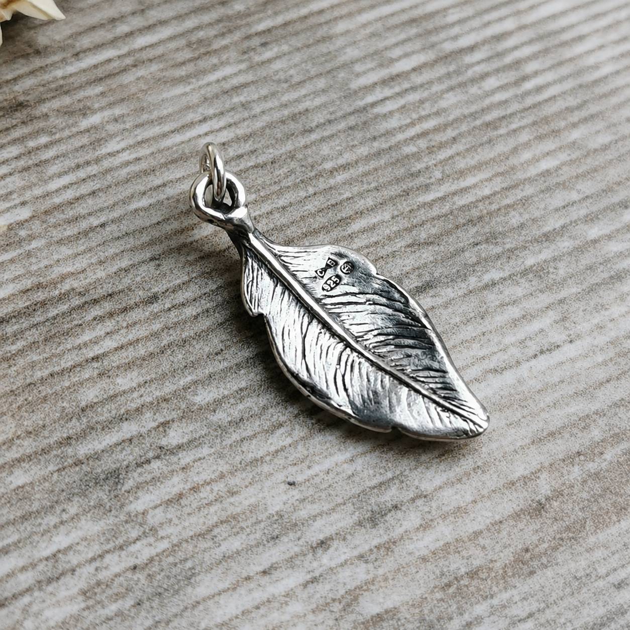 A sterling silver feather pendant with detailed textured lines and a '925' silver purity stamp and hallmark, resting on a rustic grey wood surface.