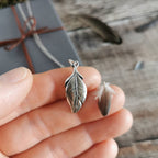 A silver feather pendant held on fingers next to a small feather, with a blurred grey gift box in the background on a rustic wooden surface.