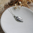 A silver feather pendant necklace on a delicate silver chain, displayed on a round white ceramic dish with dried flowers in the background.