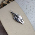 A silver feather-shaped pendant with detailed textures and a small loop at the top, displayed on a ceramic surface