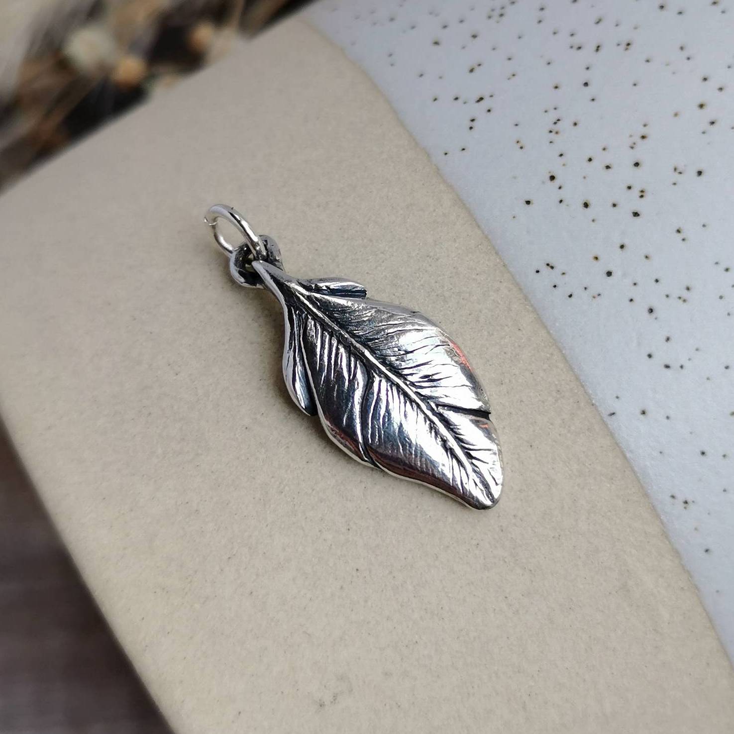 A silver feather-shaped pendant with detailed textures and a small loop at the top, displayed on a ceramic surface