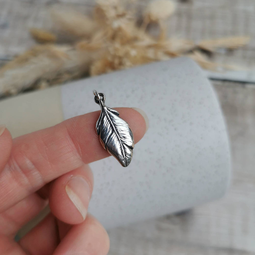 A highly detailed and dainty silver feather-shaped charm, held on a person's finger against a soft-focus background of a white textured tube and dried beige flowers.