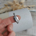 A highly detailed and dainty silver feather-shaped charm, held on a person's finger against a soft-focus background of a white textured tube and dried beige flowers.