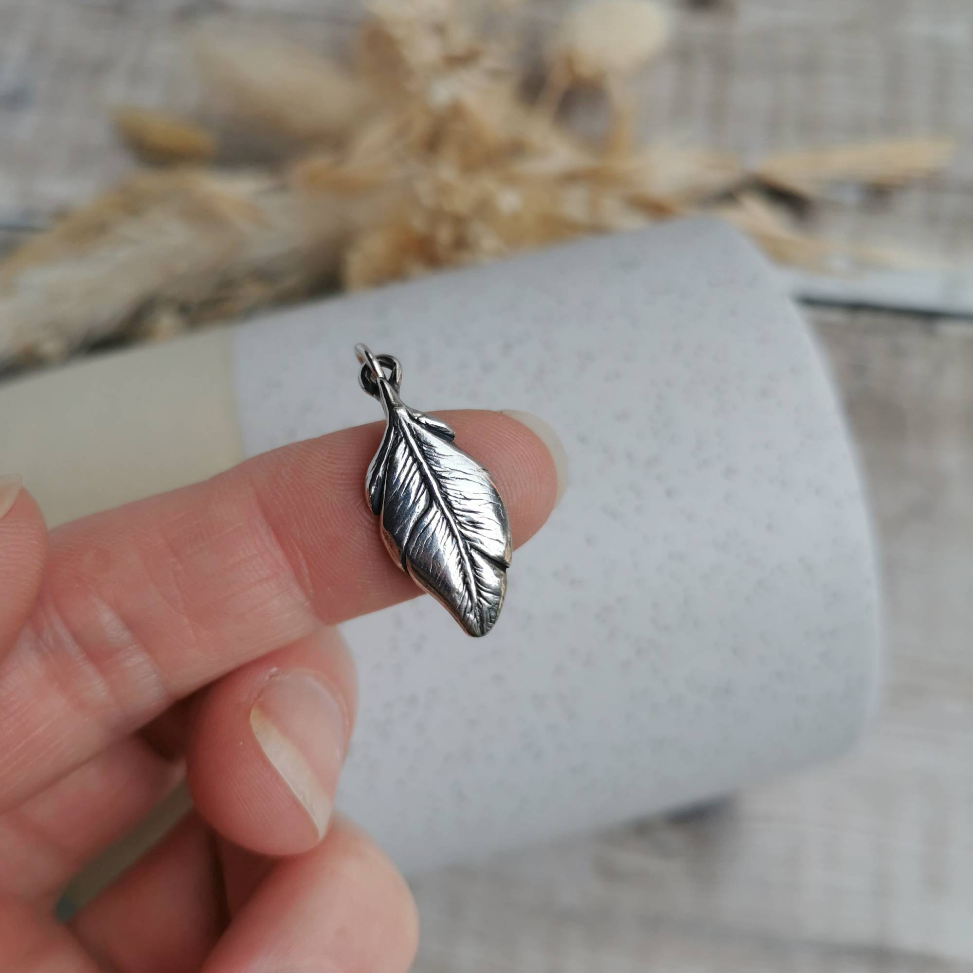 A highly detailed and dainty silver feather-shaped charm, held on a person's finger against a soft-focus background of a white textured tube and dried beige flowers.
