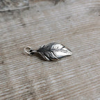 An intricately detailed silver feather charm with a small hanging loop, resting on a light brown wooden surface.