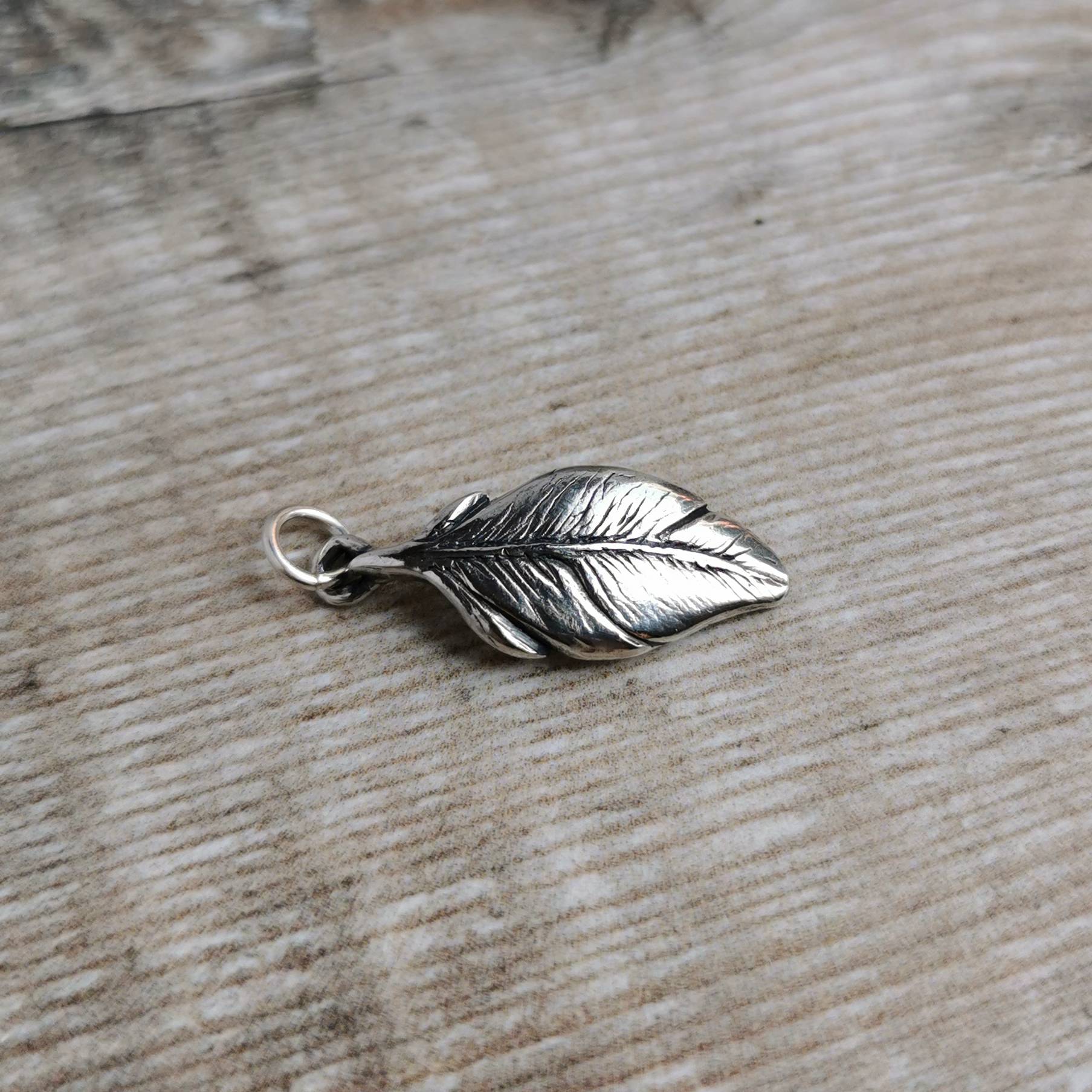 An intricately detailed silver feather charm with a small hanging loop, resting on a light brown wooden surface.
