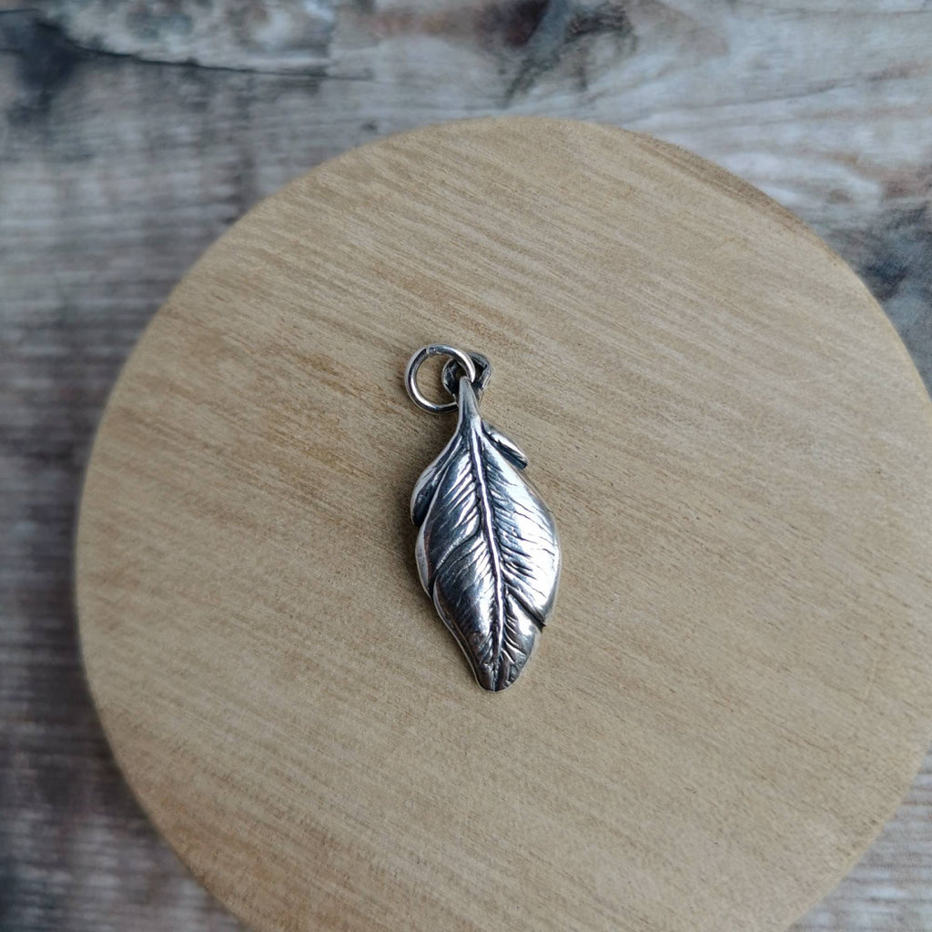A silver feather pendant charm with a small hanging loop, displayed on a round wooden coaster over a rustic wood surface.