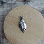 A silver feather pendant charm with a small hanging loop, displayed on a round wooden coaster over a rustic wood surface.