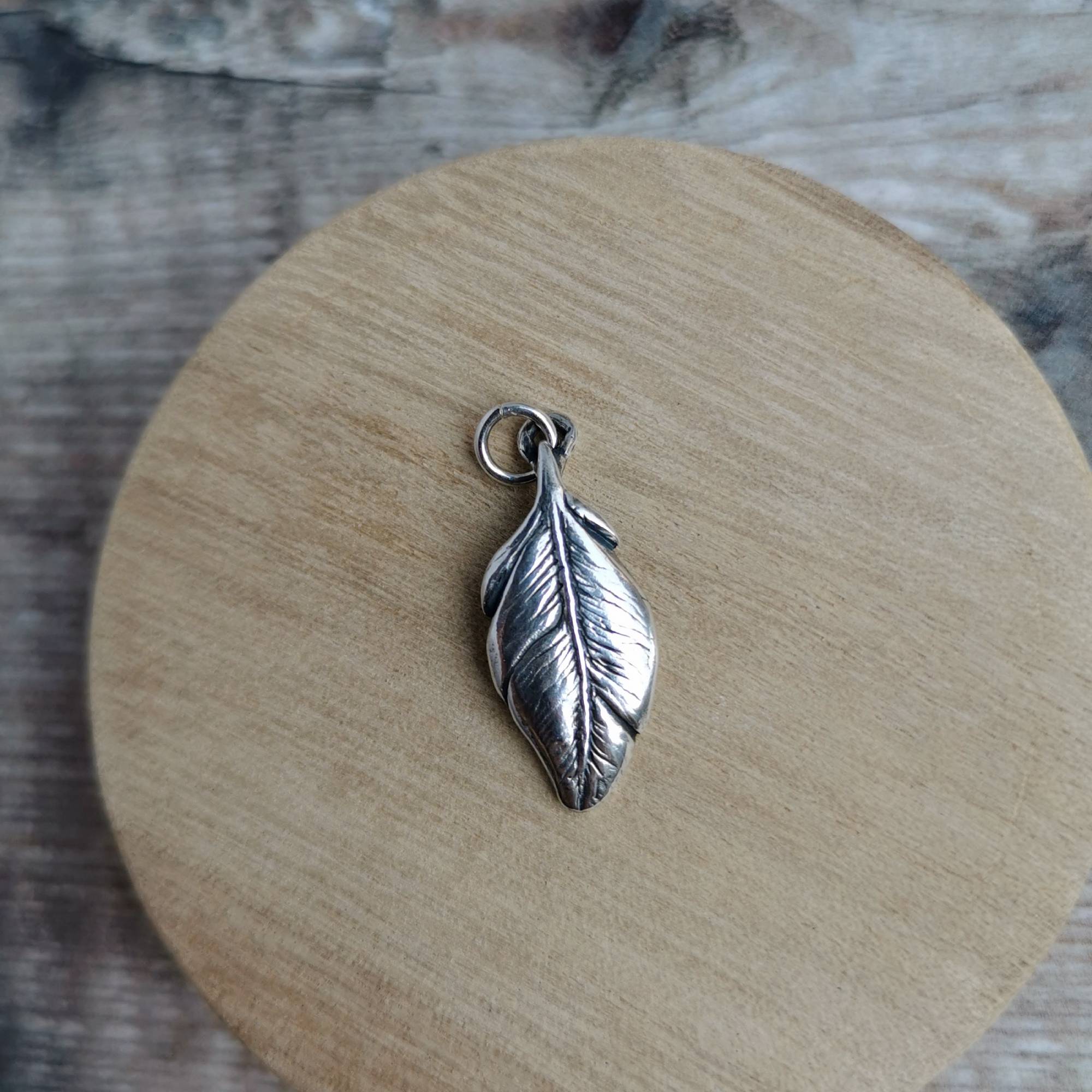 A silver feather pendant charm with a small hanging loop, displayed on a round wooden coaster over a rustic wood surface.