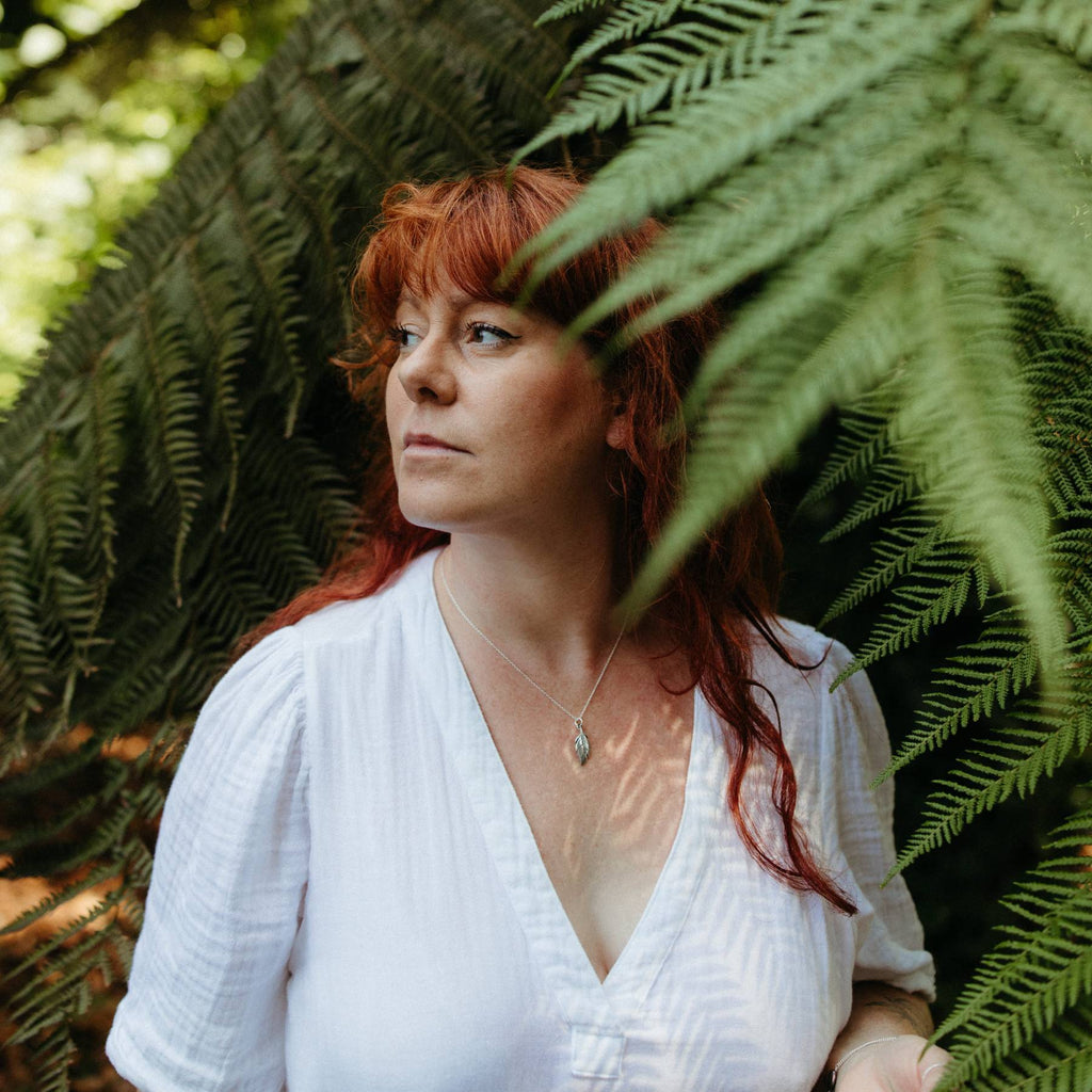 A woman with red hair wearing a white textured blouse with a silver feather charm necklace, standing in front of lush green fern leaves.