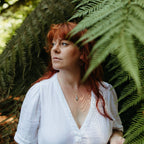 A woman with red hair wearing a white textured blouse with a silver feather charm necklace, standing in front of lush green fern leaves.
