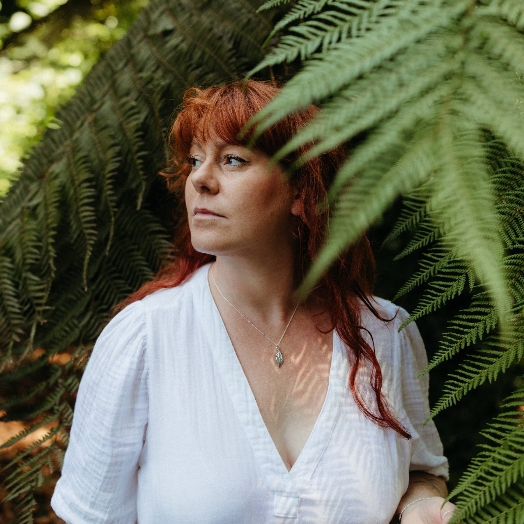 A woman with red hair wearing a white textured blouse with a silver feather charm necklace, standing in front of lush green fern leaves.