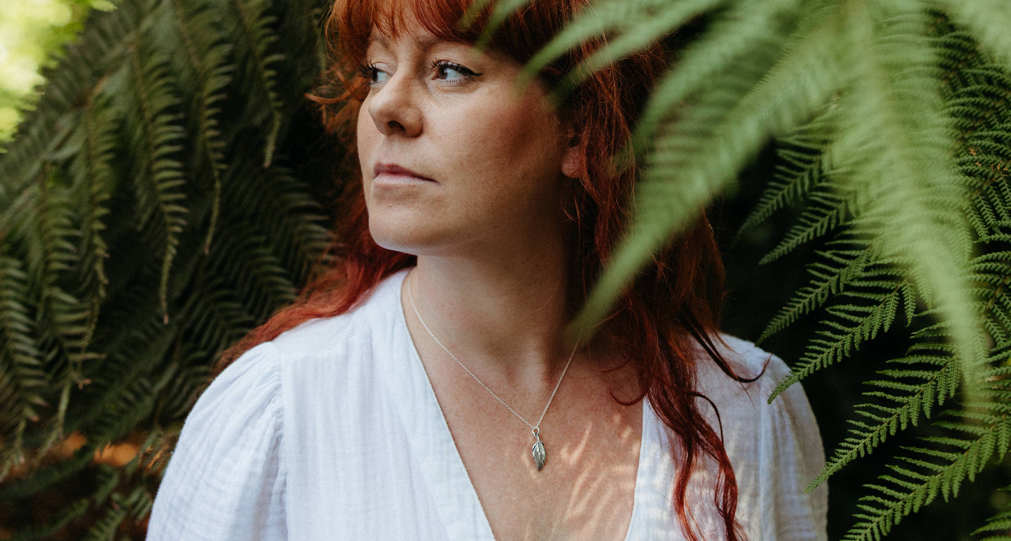 A woman with red hair wearing a white top and a silver feather pendant necklace, framed by green fern leaves.