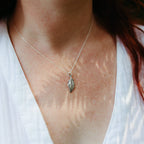 A close-up of a delicate silver chain necklace with a small feather-shaped pendant, worn by a person in a white V-neck top.