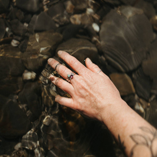 A close-up of a person's hand resting in shallow water over dark rocks, wearing multiple rings including silver bands with a small turquoise stone and an amethyst with other stacked silver rings with thumb rings and midi rings.