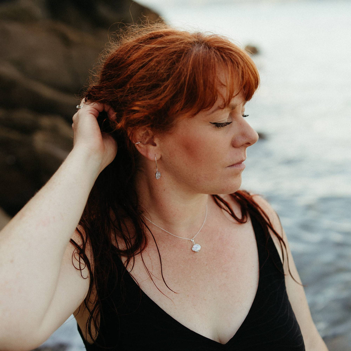 A woman with red hair wearing a black swimsuit, silver necklace matching and earrings with ivory keshi pearls, posing outdoors near water and rocks in Cornwall, UK.