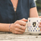 young woman holding a mug on a bench at the beach, wearing minimal handmade silver jewellery