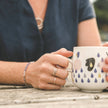 young woman holding a mug on a bench at the beach, wearing minimal handmade silver jewellery
