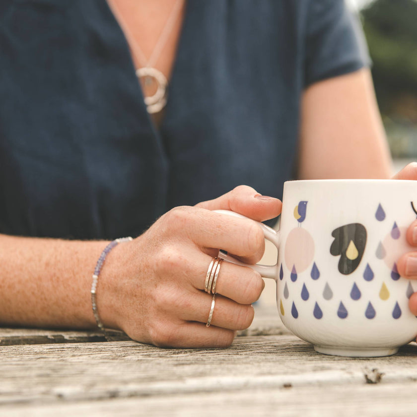 young woman holding a mug on a bench at the beach, wearing minimal handmade silver jewellery