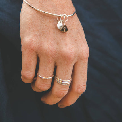 Hand wearing a solid silver bangle bracelet with charms and silver stacking rings against a navy linen dress