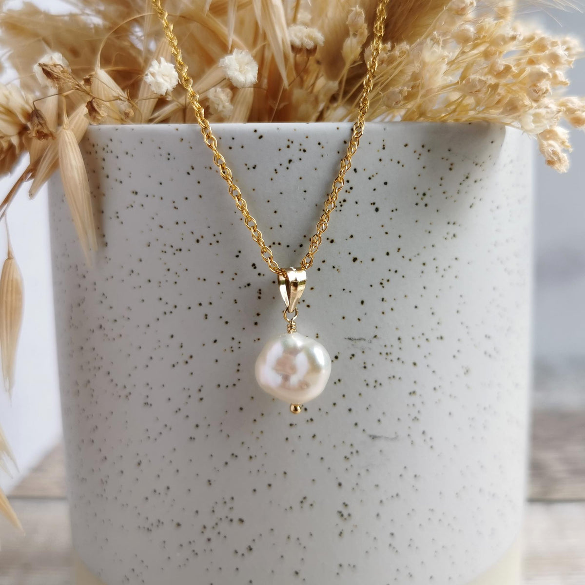 A gold chain necklace with a single irregular white freshwater keshi pearl pendant, draped over a speckled white ceramic pot filled with dried beige grasses.