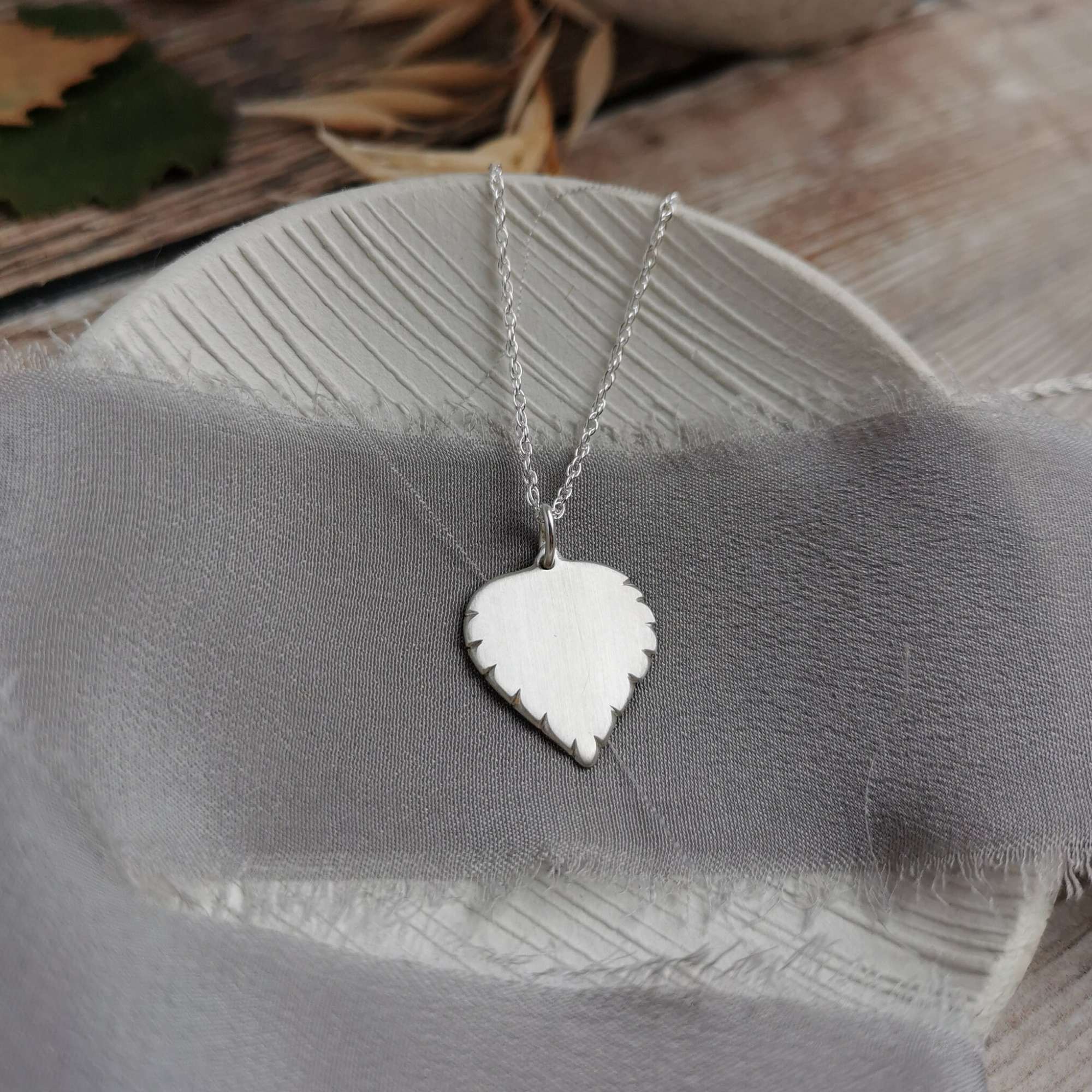 Silver birch leaf-shaped pendant necklace on a grey fabric background