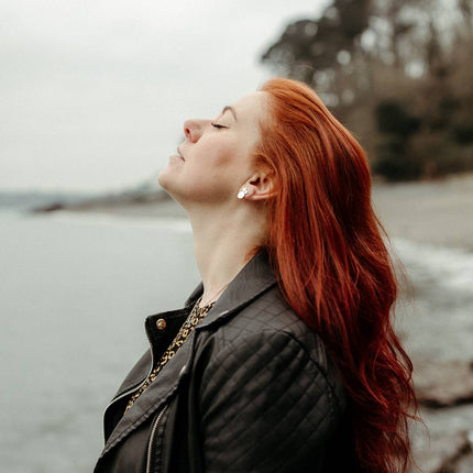 Woman with red hair wearing handmade silver mantra studs, and a black leather jacket by the sea in cornwall