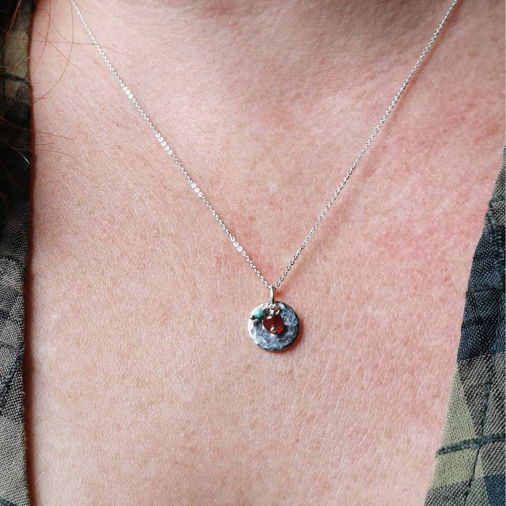 A close-up of a person's neck and chest wearing a delicate silver birthstone necklace with a small hammered silver circle featuring ruby and turquoise gemstones for july and december birth months.