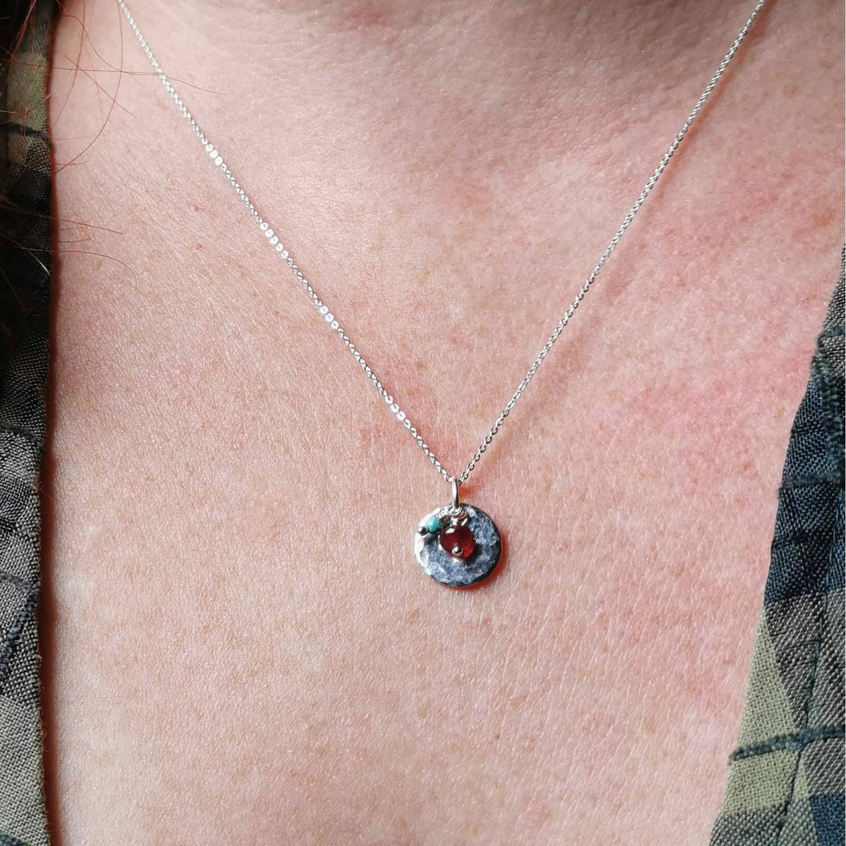 A close-up of a person's neck and chest wearing a delicate silver birthstone necklace with a small hammered silver circle featuring ruby and turquoise gemstones for july and december birth months.
