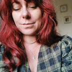 A close-up shot of a woman with vibrant red hair wearing a handmade silver birthstone necklace with a small circular hammered silver pendant with ruby and turquoise gemstone charms for july and december birthdays, worn with a green plaid shirt, in an indoor setting.