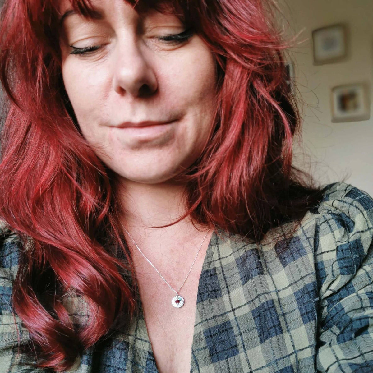 A close-up shot of a woman with vibrant red hair wearing a handmade silver birthstone necklace with a small circular hammered silver pendant with ruby and turquoise gemstone charms for july and december birthdays, worn with a green plaid shirt, in an indoor setting.