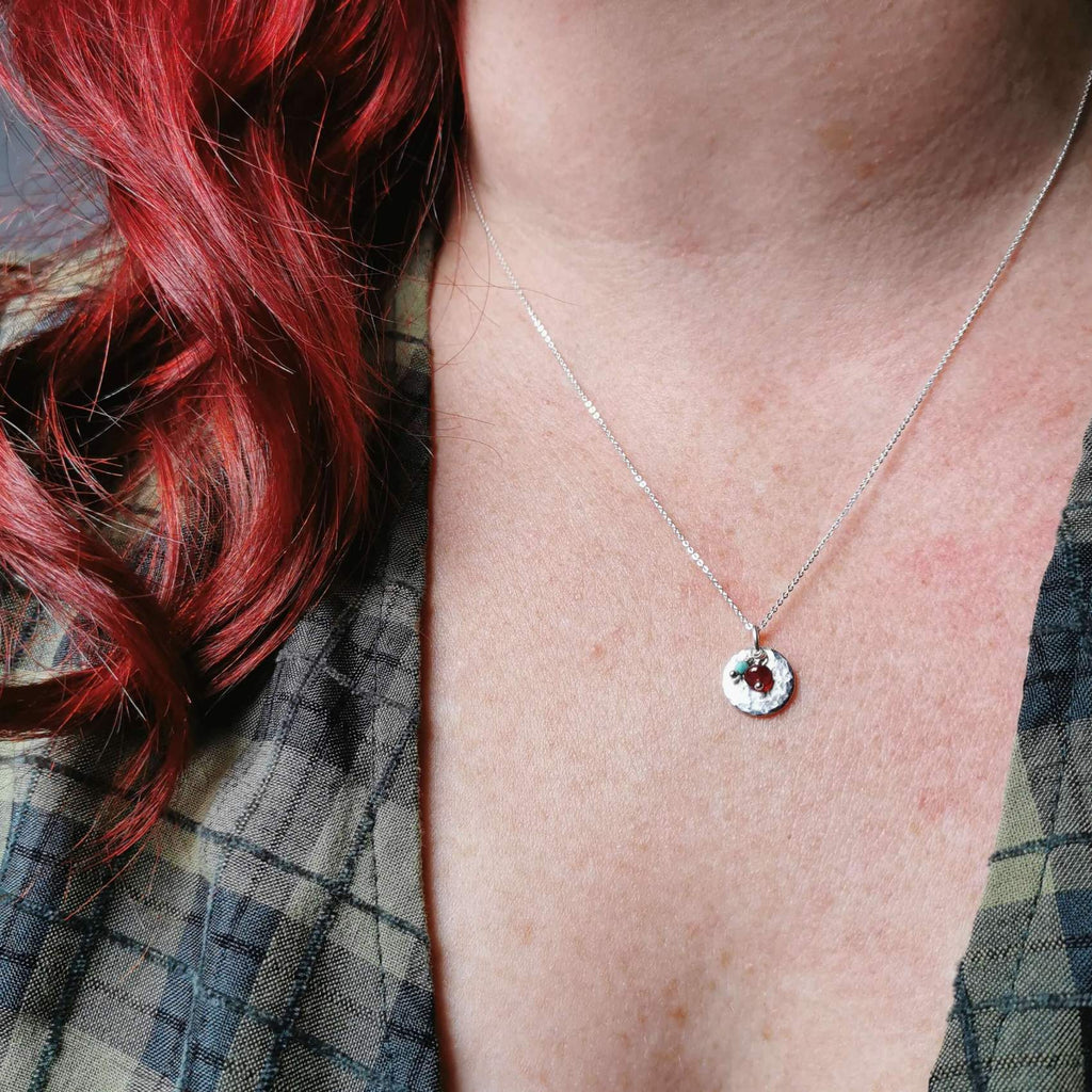 A close-up of a person wearing a delicate silver chain necklace with a small circular pendant featuring ruby and turquoise gemstone charms for july and december birthstones, styled with red hair and a plaid shirt.
